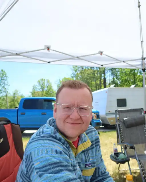 Troy sitting under a camp canopy at a campground, a pickup and trailer behind him