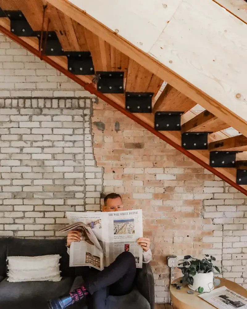 Troy reading a newspaper on a couch beneath an open wooden staircase