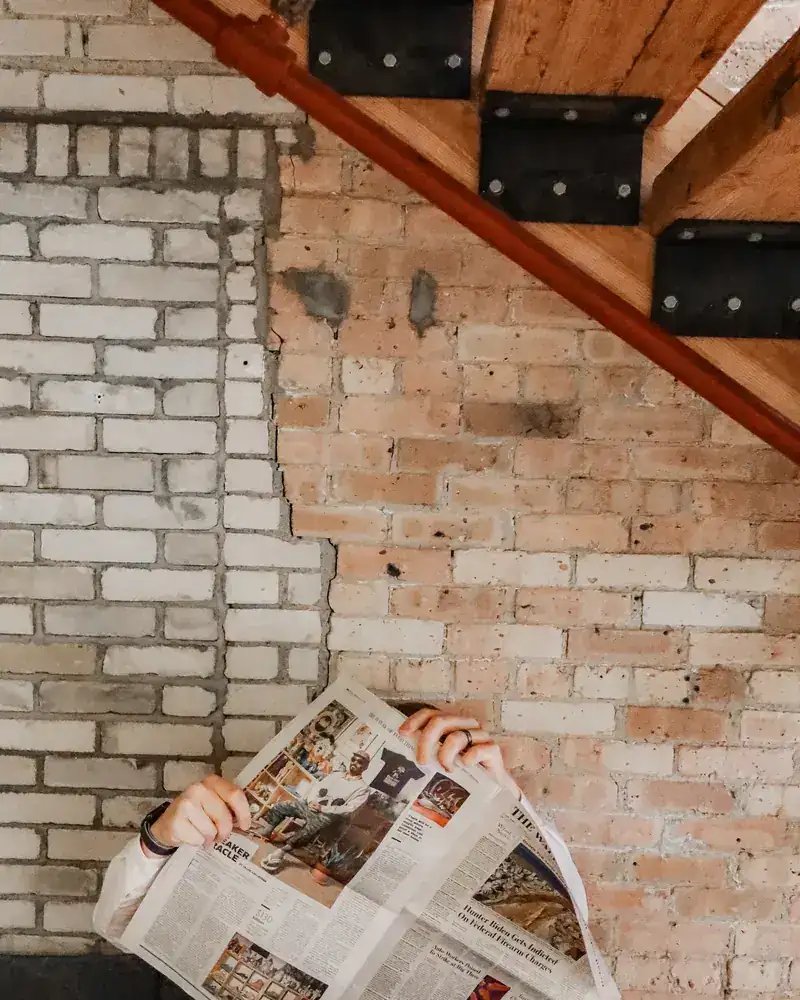 Troy hiding behind an open newspaper against a brick wall