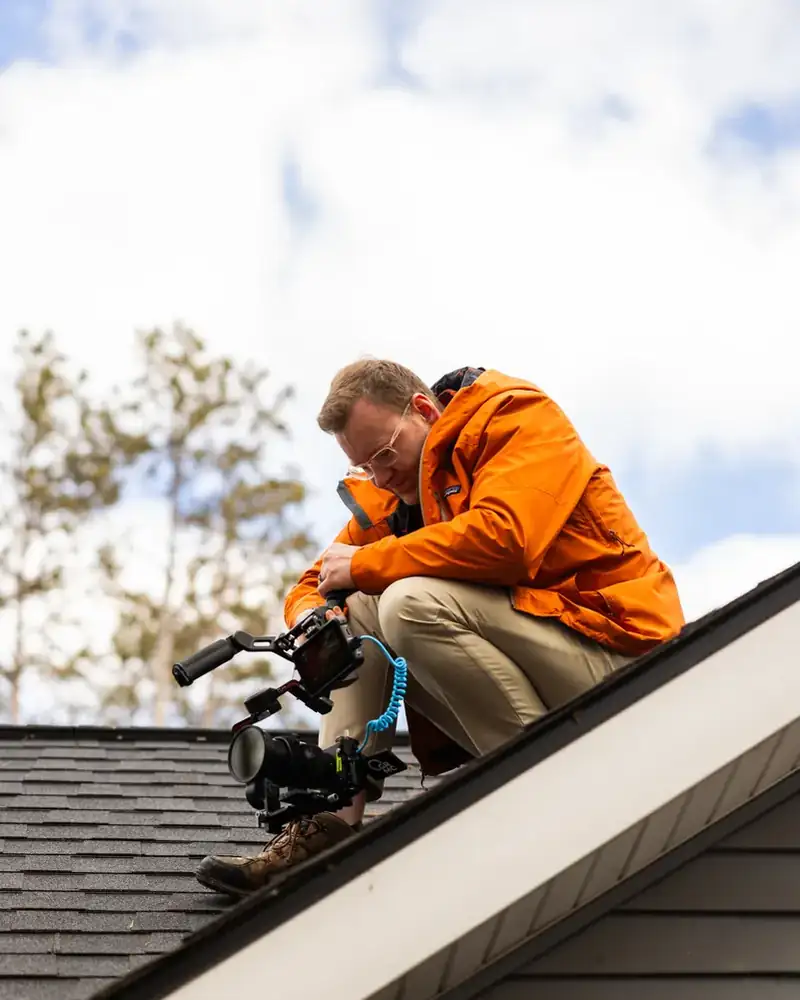 Troy crouched on a rooftop in an orange jacket, filming with a camera on a gimbal